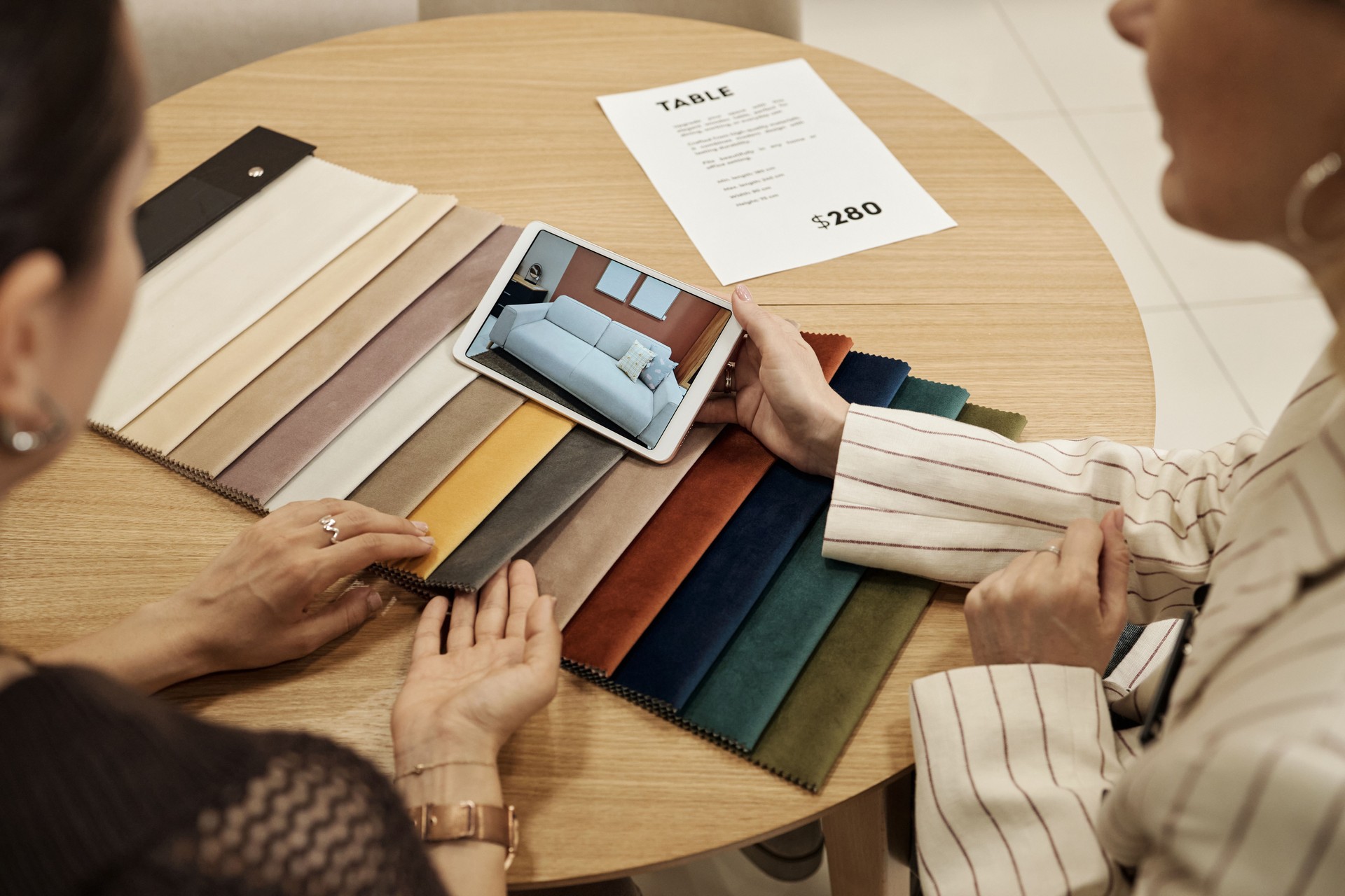 Two Young Adult Caucasian Women Discussing Sofa Fabric Samples Using Tablet