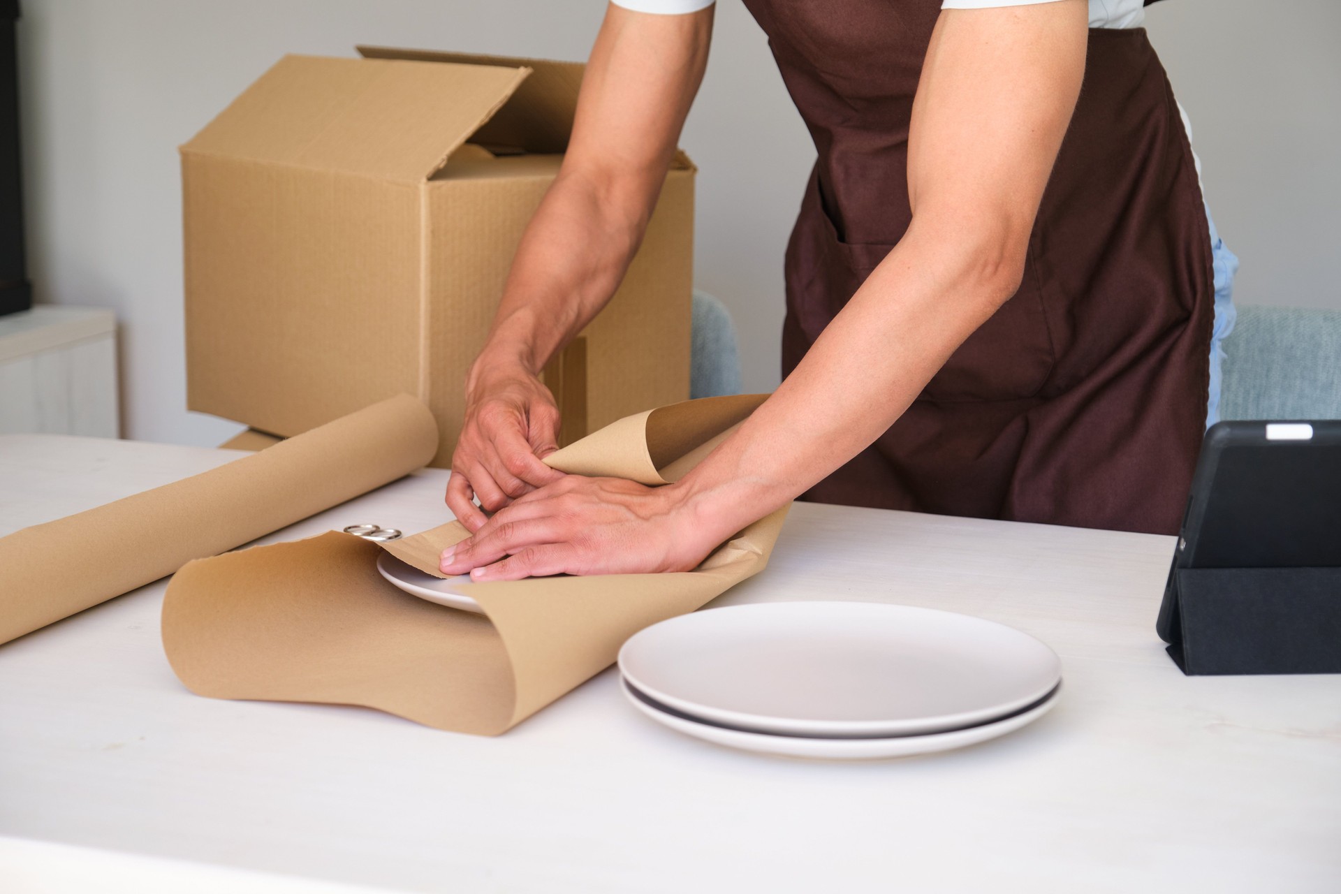 Indian man wearing brown apron packing plates using brown paper and tape at home